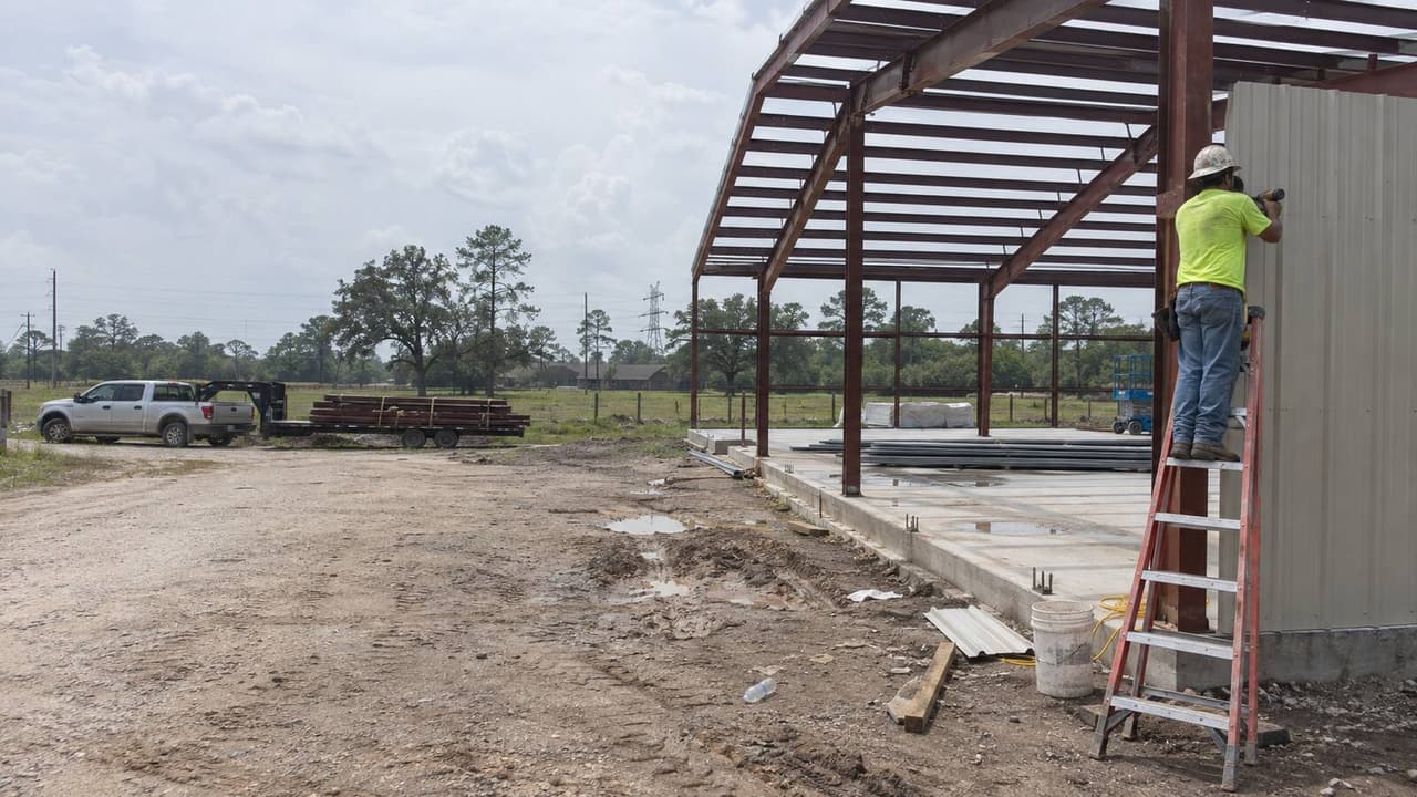 Worker sheeting the wall of a red-iron barndominium frame on acreage outside Houston